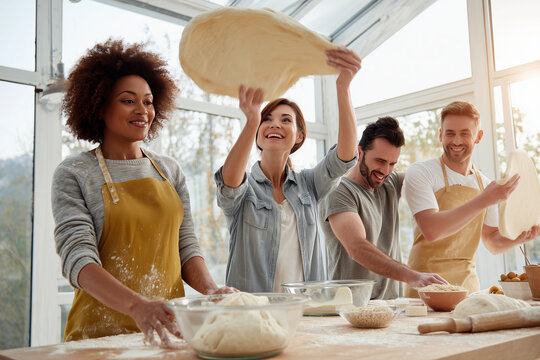 A mixed-race group of four friends joyfully stretching pizza dough high in the air in a bright kitchen with large windows suggesting cooking class, teamwork, and culinary hobby