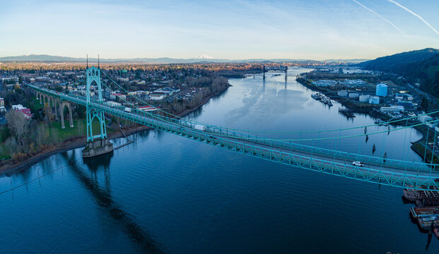 Aerial view of the iconic St. Johns Bridge casting a striking green hue against the dark, reflective Willamette River, with a backdrop of the city skyline, Portland, Oregon, United States.