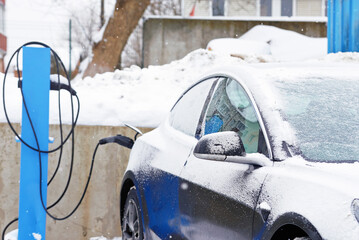 Electric car charging battery during blizzard, EV car parked at charging station and charge battery in snowy day. Electric vehicle maintenance in cold winter conditions. Selective focus