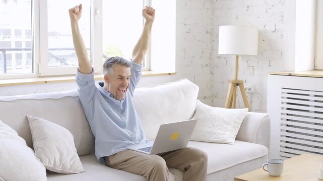 Happy man celebrating online success  or business success  using laptop while sitting on sofa at home