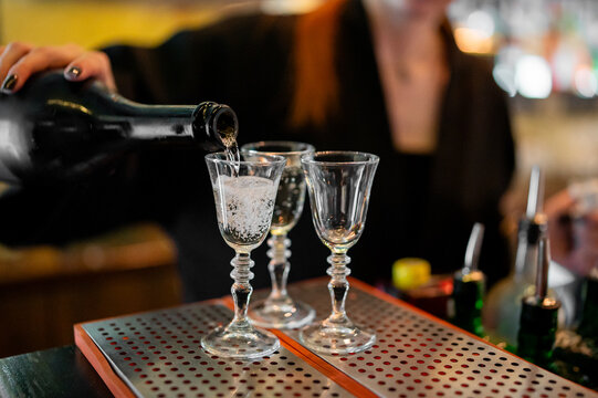 A bartender pours a sparkling clear beverage from a bottle into a row of three elegant stemmed glasses on a metallic bar top, with a dark, blurred background