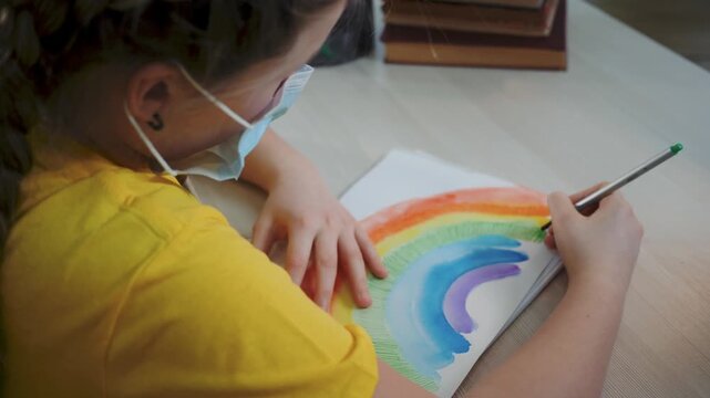 Painting rainbow while wearing mask, girl uses watercolor pencil at desk, school art project shows colorful rainbow, close up of hand and brush, child focused on creative art and education program