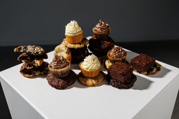Artistic arrangement of chocolate and vanilla cupcakes, stacked cookies, and fudgy brownies on a white pedestal with a gray backdrop and copy space