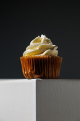 Single vanilla cupcake with smooth, swirled frosting placed at the edge of a white surface against a dark gradient background, with copy space