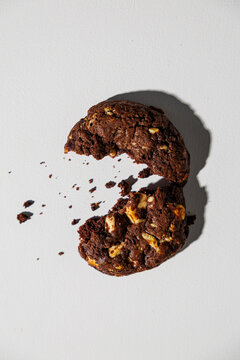 A dark chocolate cookie with visible nut chunks is split in half, with crumbs scattered across a white background, creating a dynamic and appetizing composition