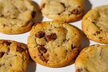 A detailed view of golden brown chocolate chip cookies arranged on a white surface, showing melty chips and soft texture under natural light. No visible branding
