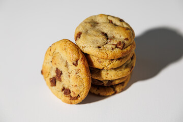 A neat stack of golden chocolate chip cookies with a single cookie leaning forward, set against a clean white and grey background. No visible logos or branding