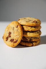 A neat stack of golden chocolate chip cookies with a single cookie leaning forward, set against a clean white and grey background. No visible logos or branding