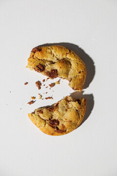 A chocolate chip cookie broken in half lies on a white surface, with scattered crumbs adding a touch of realism. No visible branding or packaging