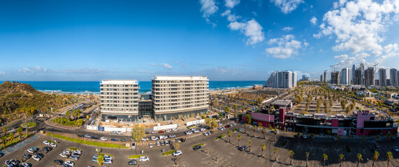 Aerial view of the coastline meeting modern buildings, a large car park, and high-rise buildings under a blue sky, Rishon LeZion, Center District, Israel.