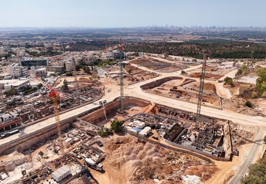 Aerial view of construction site with cranes and deep excavations under a clear sky, contrasting with the distant city skyline, El'ad, Center District, Israel.