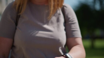 Close up of student checking time on smartwatch outdoors during bright sunny day, fingers adjusting screen under natural light, blurred green background