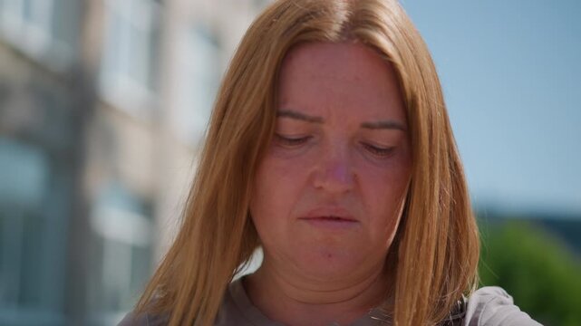 Close up of worried woman with long hair looking down thoughtfully under bright sunlight, wearing backstrap, blurred residential building background