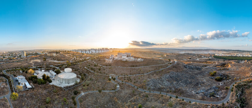 Aerial view of golden light washes over the arid landscape, illuminating the modern structures and winding roads of Migdal Tsedek, Rosh Haayin, Israel.