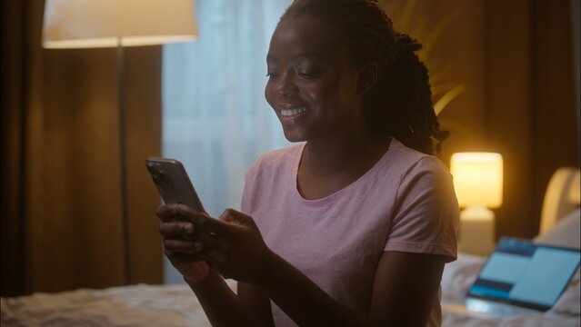 Cheerful glad African American ethnic business woman female girl businesswoman using mobile phone thinking answer texting dating online app sending message SMS work sitting on bed laptop at home night
