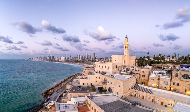 Aerial view of ancient St. Peter's Church stone buildings meet the turquoise sea, contrasting with the modern skyline under a pastel sky, Old Jaffa, Tel Aviv-Yafo, Israel.