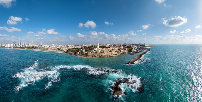 Aerial view of the ancient port city, where the turquoise sea crashes against the rugged coastline under a vast, cloud-dotted sky, Old Jaffa, Tel Aviv-Yafo, Israel.
