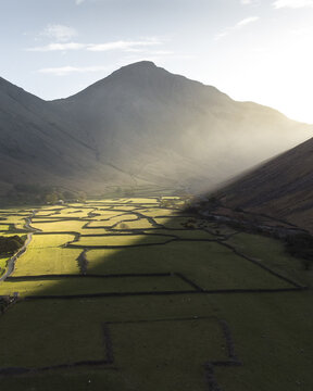 Aerial view of sun-drenched fields at Wast Water bordered by dark stone walls nestled in a valley beneath towering peaks, a contrast of light and shadow, Cumbria, England, United Kingdom.