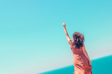 A young girl in a summer brown dress with a raised hand enjoys a vacation at the sea