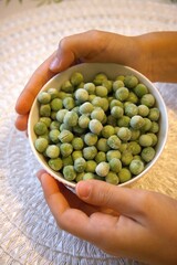 Child holding a white bowl filled with frozen green pea, photographed in natural condition. Healthy eating and childhood nutrition concept.
