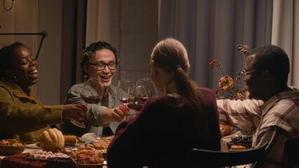 Stab shot of four diverse young people clinking wine glasses while celebrating Friendsgiving sitting at festive table with homemade food, pumpkins and snacks
