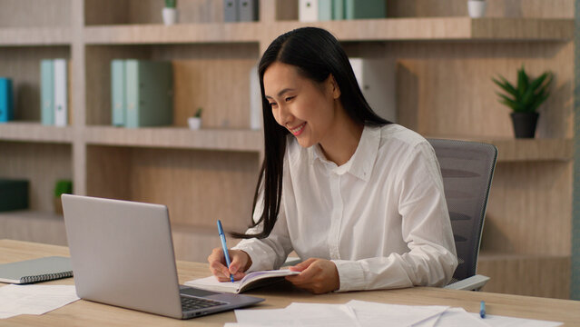 Happy smiling Chinese businesswoman Asian girl worker Korean Japanese business woman female secretary manager online laptop desktop call remote conference meeting writing notes pen notebook in office