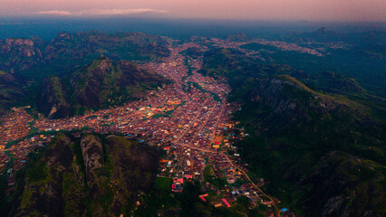 Aerial view of a sprawling town nestled among rugged, verdant hills under a dusky sky, the buildings a mosaic of warm hues against the cool green landscape, Idanre, Ondo, Nigeria.