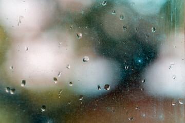 Macro close-up of raindrops on a dusty window. The outside world is blurred into a soft, abstract bokeh of green, white, and blue colors.
