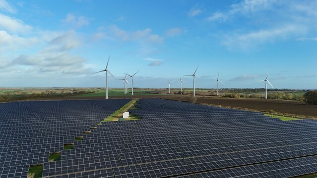 Aerial view of solar panels reflecting the sky with wind turbines in the background under a blue sky with scattered clouds, Mansfield, England, United Kingdom.