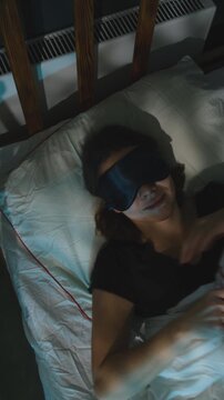 Vertical video. Woman lying in bed preparing for healthy deep sleep by inserting earplugs in her ears and wearing a sleep mask