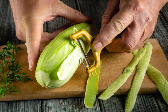 A person uses a peeler to remove the skin from a green vegetable while sitting at a wooden cutting board. Fresh green peelings are visible on the surface next to the vegetable