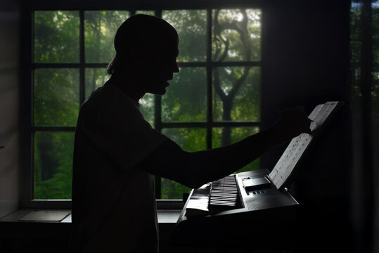 Silhouette of man playing synthesizer at home by his window. Silhouette of man writing music notes at keyboard with forest view