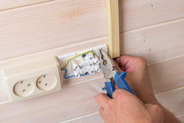 Close up of electrician hands with pliers as he tightens copper conductor into outlet terminal on pale wood paneling, detail focus.