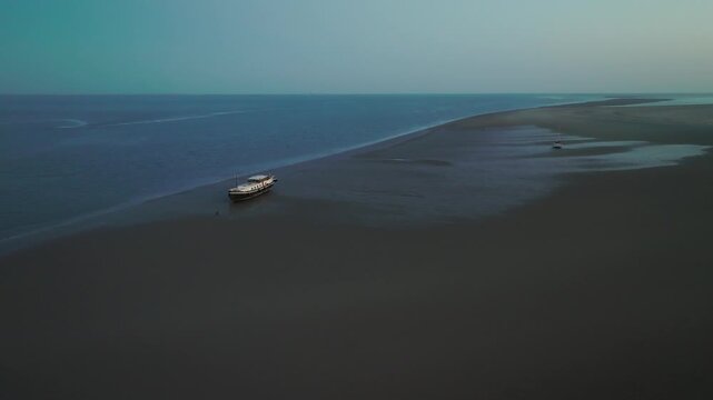 Classic luxemotor rests on a narrow sandbar during blue hour; drone makes a smooth forward glide along the shoreline over dark intertidal flats. Wadden Sea.