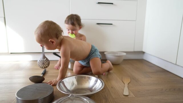 Toddler and baby play with cookware on wooden kitchen floor surrounded by metal pot pan bowl spoon and wooden spatula toddler reaches for pan while sibling sits behind holding silicone teether