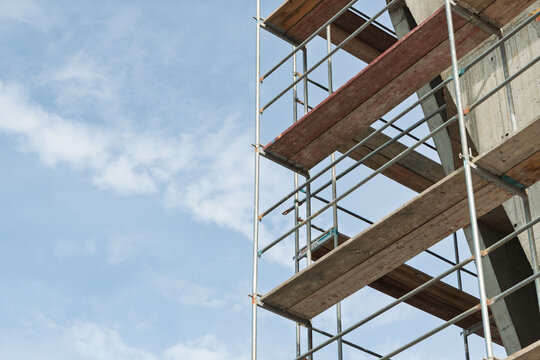 Close view of metal scaffolding and planks on concrete frame, vertical pipes and guardrails in geometric arrangement against sky.