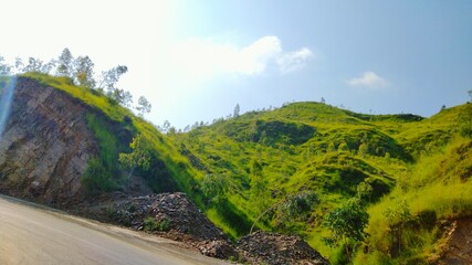 Lush green rolling hills under a bright blue sky, with a rocky hillside and a road.