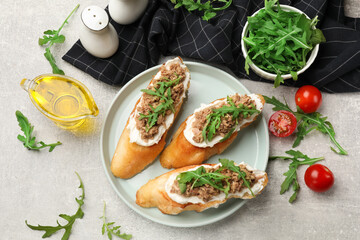 Delicious bruschettas with tuna, tomatoes, oil and arugula on grey table, flat lay