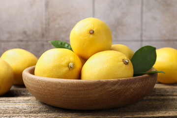 Fresh lemons with green leaves and bowl on wooden table near tiled wall, closeup