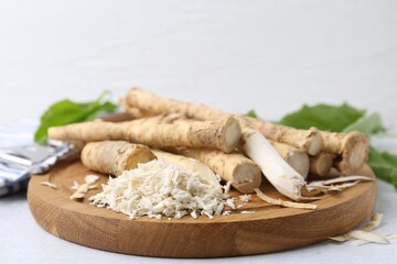 Whole and grated horseradish roots, peeler and green leaves on light grey table, closeup