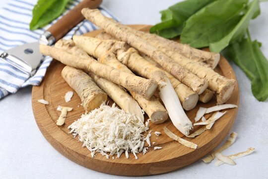 Whole and grated horseradish roots, peeler and green leaves on light grey table, closeup