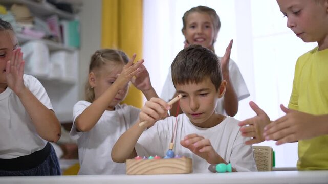 Boy lights candle on cake while child and friend clap and smile around birthday toy in classroom during celebration with playful gesture curious expression and hands reaching toward candle joyful - Powered by Adobe