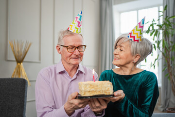 Make a wish. Family senior couple in party hat cap celebrating birthday anniversary together at home. Old man blowing out burning candles on birthday cake. Old woman wishes husband happy birthday