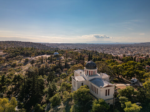 Aerial view of the National Observatory of Athens standing proudly amidst verdant trees, contrasting with the distant cityscape under a serene sky, Athens, Central Athens Regional Unit, Greece.