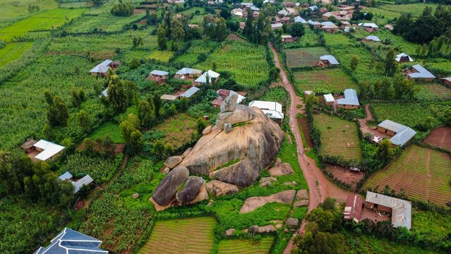 Aerial view of a colossal rock formation amidst verdant fields and scattered dwellings, a striking contrast of earth tones and vibrant greens, Riyom, Plateau, Nigeria.