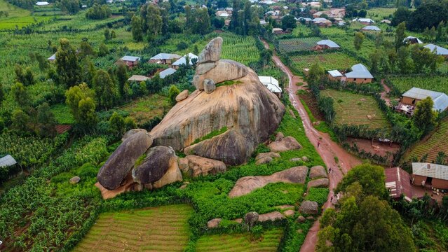 Aerial view of a colossal rock formation surrounded by verdant fields, with houses nestled among the trees, creating a striking contrast of natural and built environments, Riyom, Plateau, Nigeria.