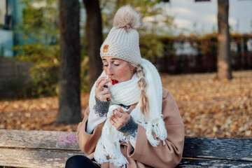 Woman bundled in coat using nasal spray outdoors autumn leaves falling around her easing cold...