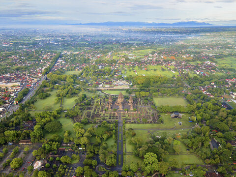 Aerial view of the majestic Prambanan Temple complex amidst lush greenery, framed by the cityscape's soft hues, Yogyakarta, Daerah Istimewa Yogyakarta, Indonesia.