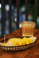 A serving of Pisang Goreng Keju (fried bananas with shredded cheese) in a basket, paired with a glass of iced coffee latte on a wooden table in a cafe with warm lighting.