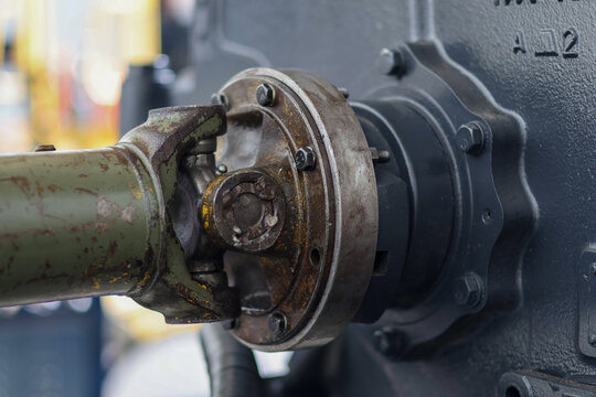 Close-up of a greasy, worn universal joint and driveshaft coupling on a heavy machinery transmission, showing rust, wear, and an attached hydraulic hose.
